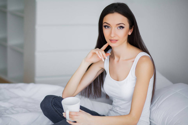Goedemorgen. Portret van een lachende ontspannen in witte bed mooie jonge brunette vrouw - Foto, afbeelding