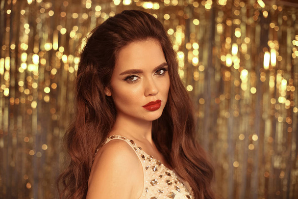 Beauty Fashion Girl Portrait Isolated on golden Christmas glitter lights bokeh Background. Glamour Makeup. Gold Jewelry. Hairstyle. Alluring brunette with sensual red lipstick looking at camera. - 写真・画像