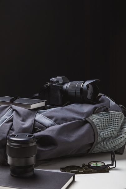 rugzak, fotocamera met lens, notebooks met pen en een kompas op zwart, reizen concept - Foto, afbeelding
