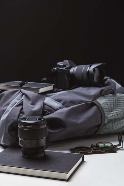 sac à dos, appareil photo avec objectif, cahiers avec stylo et boussole sur noir
   - Photo, image