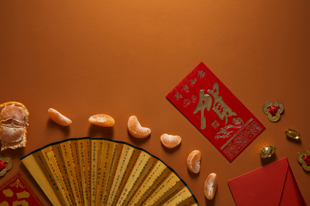 top view of peeled tangerine, fan with hieroglyphs and red envelope on brown background - Photo, image