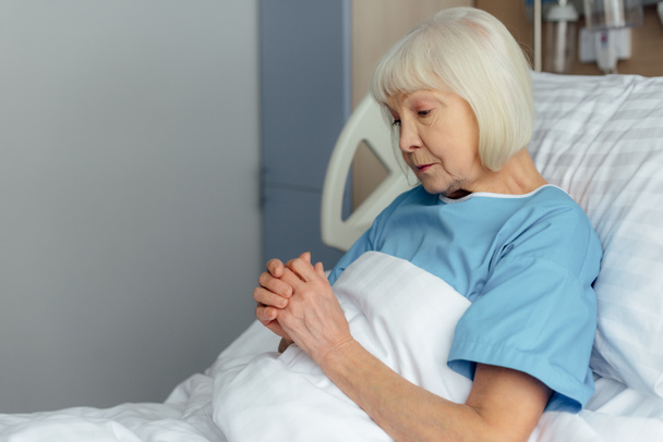 selektywne focus starszy Kobieta, leżąc w łóżku z założonymi rękoma i modląc się w szpitalu  - Zdjęcie, obraz