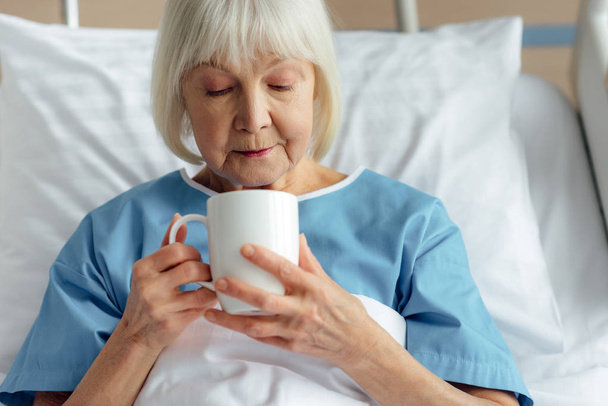 senior woman with grey hair lying in bed and drinking tea in hospital - Foto, Imagen