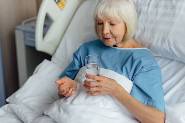 senior woman lying in bed and holding pills with glass of water in hospital - Photo, Image