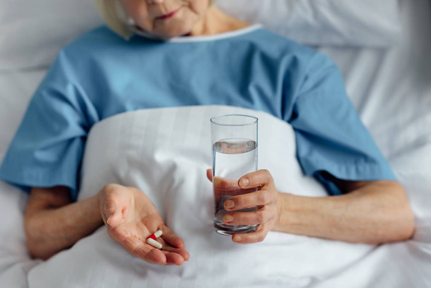 частичный взгляд пожилой женщины лежащей в постели и держащей таблетки со стаканом воды в больнице
 - Фото, изображение