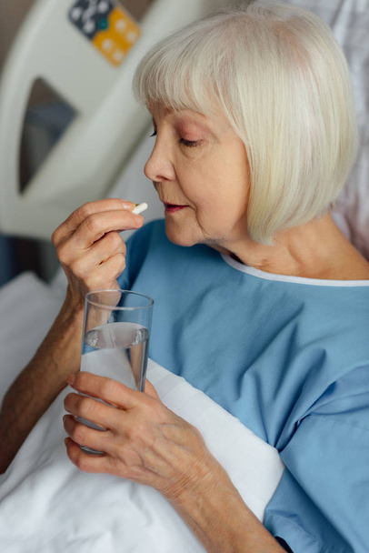 mujer mayor acostada en la cama, sosteniendo un vaso de agua y tomando pastillas en el hospital
 - Foto, Imagen