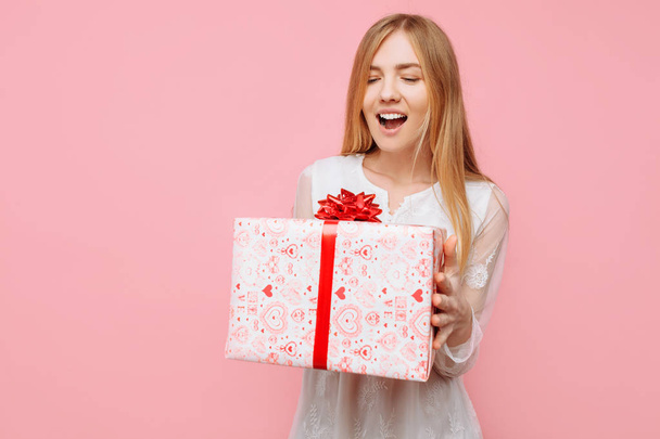 Portrait of a happy young woman in a white dress, with a gift box in her hands, delighted with a gift - Фото, изображение