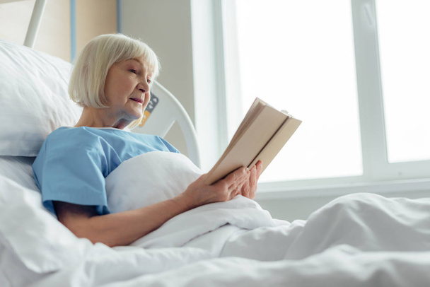senior woman with grey hair lying in bed and reading book in hospital - Photo, image