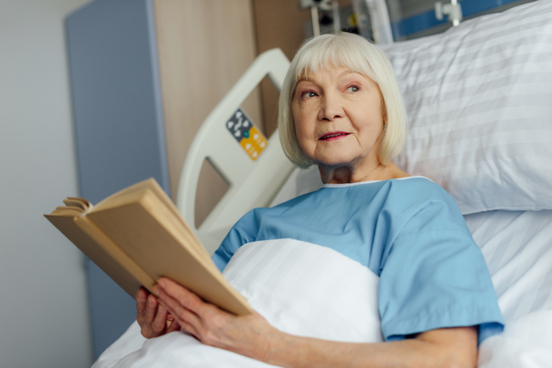 Senior vrouw met grijze haren liggend in bed en het lezen boek in ziekenhuis - Foto, afbeelding