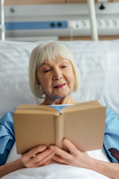 yatakta yatan ve hastanede kitap okuma gri saçlı kadın kıdemli seçici odak - Fotoğraf, Görsel