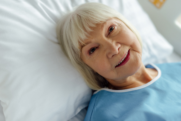Selektivní fokus usměvavá starší ženy s šedými vlasy, ležící v posteli a v nemocnici - Fotografie, Obrázek