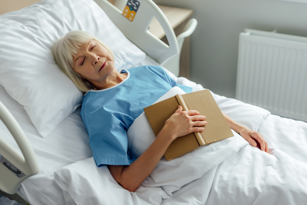selective focus of senior woman with book sleeping in bed in hospital - 写真・画像