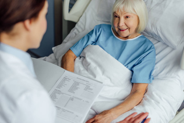 医師診断を押しながら病院のベッドで横になっている年配の女性の笑顔をコンサルティング  - 写真・画像