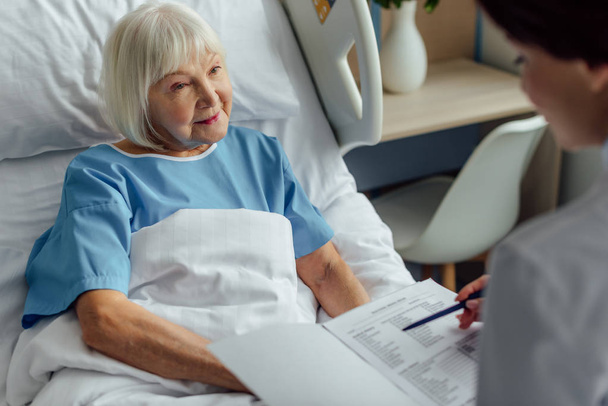 lekarz trzymając diagnozy i konsultacji starszy kobieta z siwymi włosami, leżącego w łóżku szpitalnym  - Zdjęcie, obraz