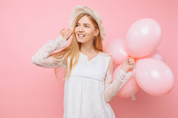 Happy girl in a white dress, in a straw hat, holds balloons in her hands, on a pink background - Foto, Imagen