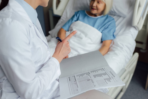 vista recortada de la doctora sentada en la cama, sosteniendo el diagnóstico y señalando con el dedo mientras la mujer mayor yacía en la cama en el hospital
 - Foto, imagen