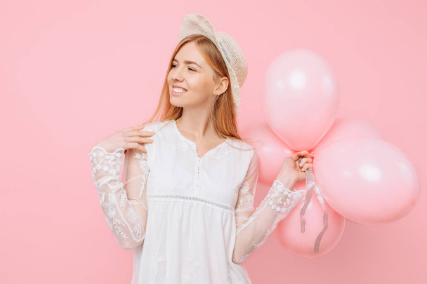 Happy girl in a white dress, in a straw hat, holds balloons in her hands, on a pink background - Фото, зображення
