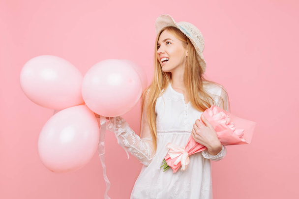 A pensive girl in a white dress, in a straw hat, stands with a bouquet of pink tulips and balloons, on a pink background. March 8 concept - Photo, image