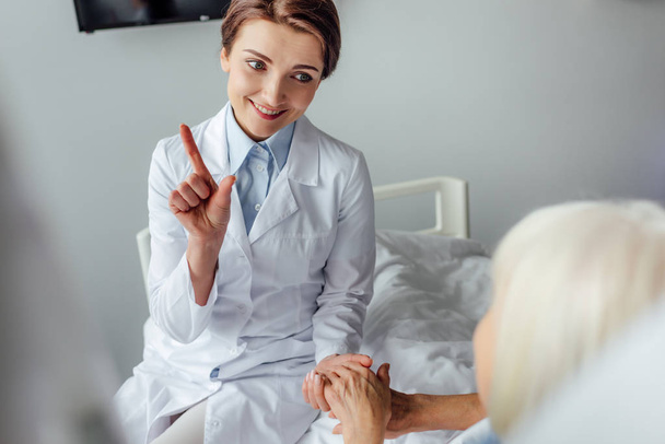 hermosa sonriente doctora señalando con el dedo y tomándose de la mano con la mujer mayor acostada en la cama en el hospital
 - Foto, Imagen