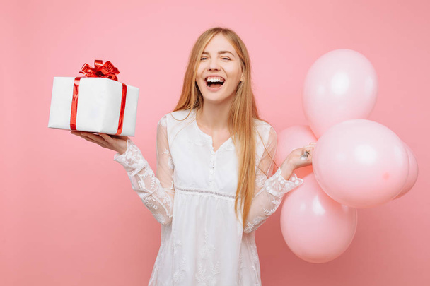 Enthusiastic young woman, in a white dress, with a gift box and balloons in their hands, on a pink background - Foto, afbeelding