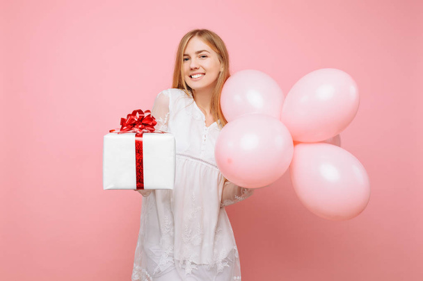 Happy young woman, in a white dress, with a gift box and balloons in their hands, on a pink background - Photo, image