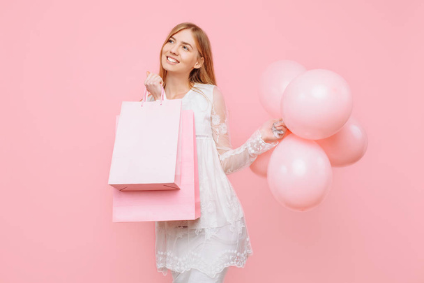 Happy young woman, in a white dress, with bags in hands and balloons, on a pink background - Foto, Imagem