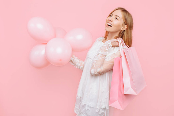 Happy young woman, in a white dress, with bags in hands and balloons, on a pink background - Fotó, kép