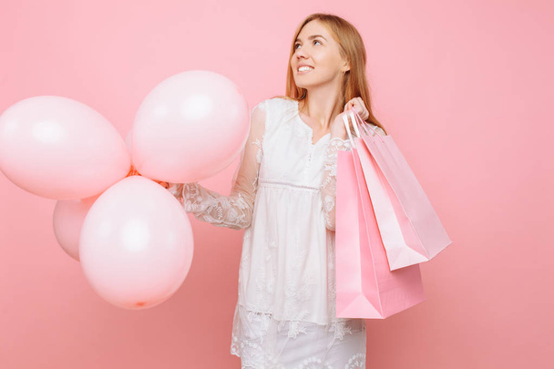 Pensive young woman, in a white dress, with bags in hands and balloons, on a pink background - Foto, Imagen