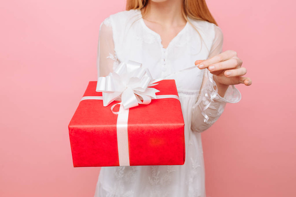 A curious girl in a white dress with a gift box in her hand, wants to see what is in the box, on a pink background - Foto, imagen