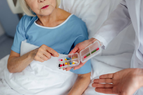 обрезанный вид женщины-врача, дающей таблетки пожилой женщине, лежащей на больничной койке
 - Фото, изображение