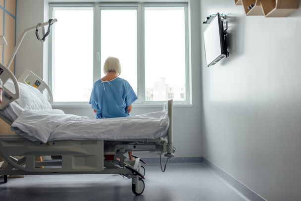 Вид сзади одинокой пожилой женщины, сидящей на кровати в больничной палате с пространством для копирования
 - Фото, изображение