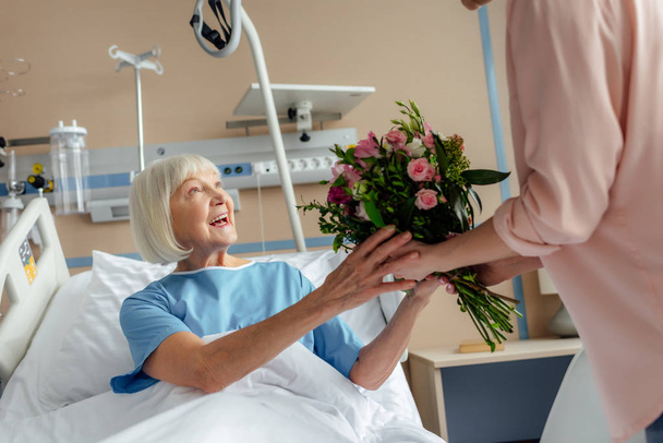 Κόρη παρουσιάζοντας λουλούδια χαμογελαστό ανώτερων γυναίκα ξαπλωμένη στο κρεβάτι στο νοσοκομείο - Φωτογραφία, εικόνα