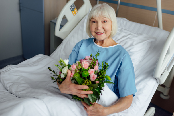 mutlu çiçek ile yatakta yalan ve kamera hastanede bakarak kadın kıdemli kurtarma  - Fotoğraf, Görsel
