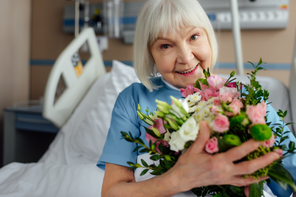 happy recovering senior woman on bed with flowers in hospital - Фото, изображение