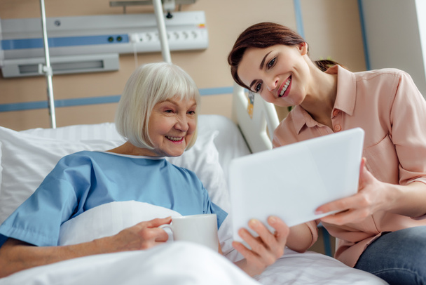 Glückliche Seniorin und Tochter mit digitalem Tablet im Krankenhausbett - Foto, Bild