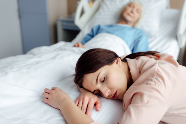 selectieve aandacht van moe slapende dochter met zieke moeder in bed op achtergrond in het ziekenhuis - Foto, afbeelding