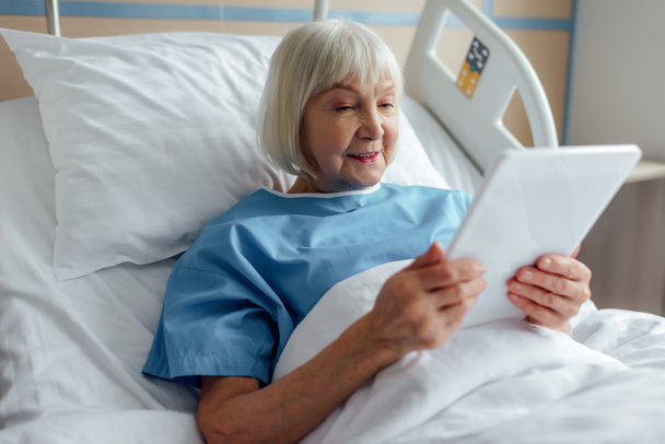 hymyilevä vanhempi nainen makaa sängyssä ja käyttää digitaalista tablettia sairaalassa
 - Valokuva, kuva