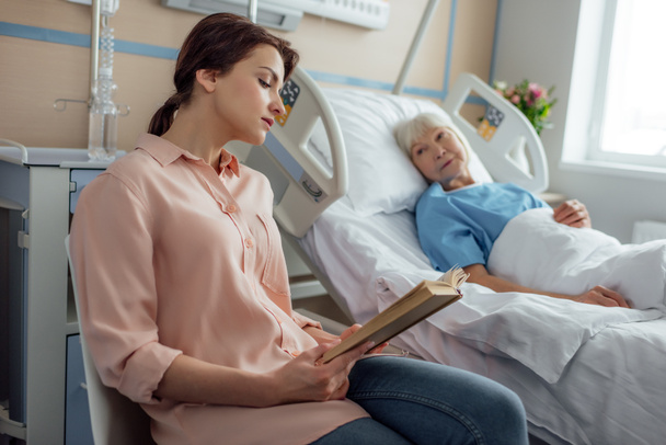 beautiful daughter reading book to sick senior mother lying in bed in hospital - Photo, Image