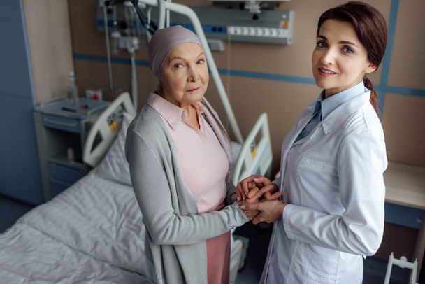 female doctor looking at camera and holding hands with senior woman in kerchief with cancer in hospital - Photo, Image