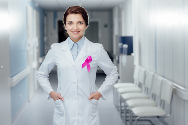 smiling female doctor with pink ribbon and hands akimbo in hospital, breast cancer awareness concept - Photo, Image