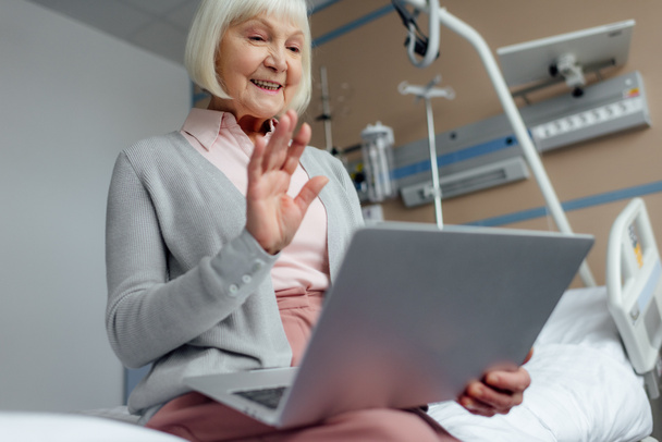 glückliche Seniorin sitzt auf dem Bett, benutzt Laptop und winkt mit der Hand, während sie Videoanruf im Krankenhaus hat - Foto, Bild