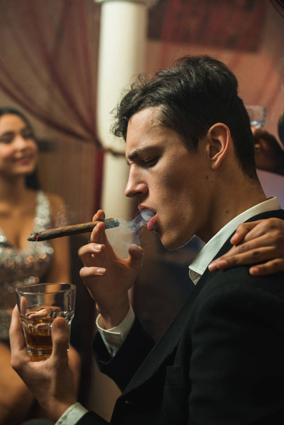Κομψό Καυκάσιος νεαρός σε μαύρο κοστούμι καπνίζοντας πούρο και πίνοντας ουίσκι το βράδυ σε μια εταιρεία με αρκετά Μέσης Ανατολής κορίτσι - Φωτογραφία, εικόνα