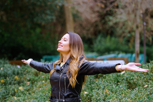 Side view of happy young woman keeping eyes closed and smiling while standing on blurred background of park trees - Photo, Image