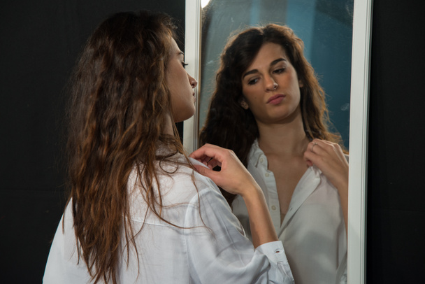 Young girl with long wavy brown hair, wearing a white men's shirt. Concept of mirroring in the early morning. Half-length portrait in a mirror. - Photo, Image