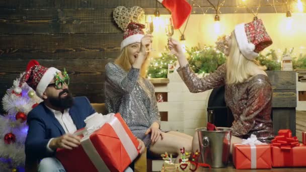 Two girls in New Years hats and brilliant silver dresses along with a bearded man who holds a large gift box drink champagne and celebrates Christmas and New Year. New Years holiday mood. - Πλάνα, βίντεο