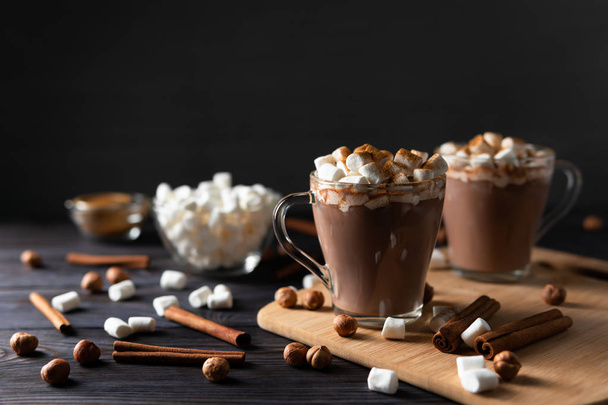 Átlátszó csésze forró csokoládé és finom édes cukorka, a fekete háttér szórás fahéj és dió közelében vágódeszka - Fotó, kép