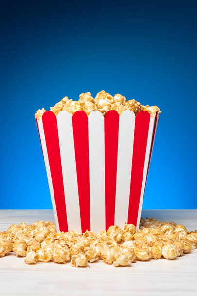 Paper bucket in red and white stripes filled with golden crunchy popcorn on blue background - Foto, afbeelding