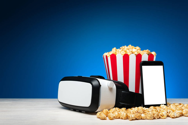 Uspořádání moderních sluchátek virtuální reality s smartphone a barevný kyblík popcornu na modrém pozadí - Fotografie, Obrázek