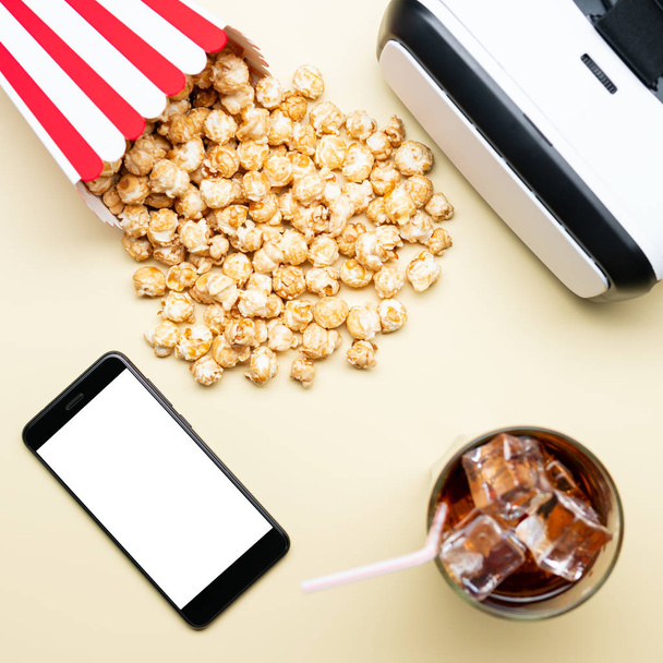 Стекло с холодным напитком и коробка свежего попкорна рядом с современными VR очками и смартфоном с чистым экраном на светло-желтом фоне
 - Фото, изображение