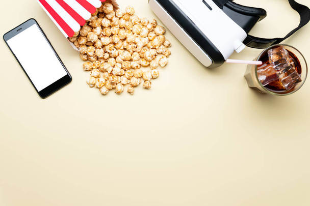 Lasi kylmä juoma ja kartonki tuoretta popcornia lähellä moderni VR suojalasit ja älypuhelin tyhjä näyttö vaaleankeltainen tausta
 - Valokuva, kuva